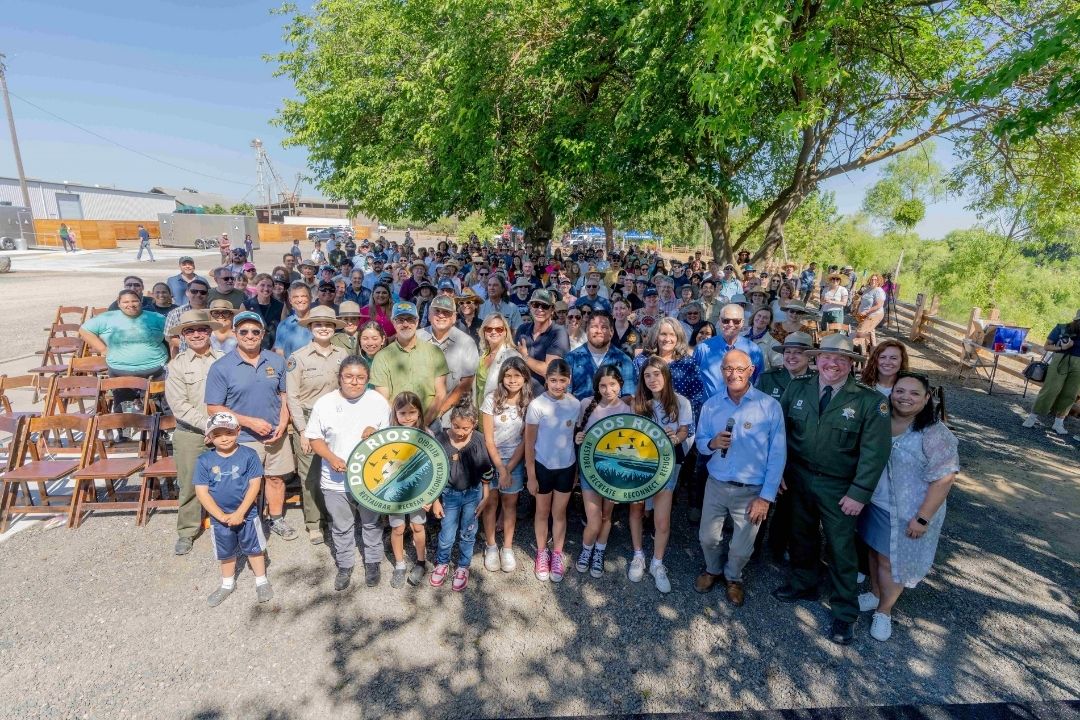 A large group of people gather outdoors, standing under a tree. The crowd includes a diverse group of individuals wearing uniforms, hats, and casual clothing.
